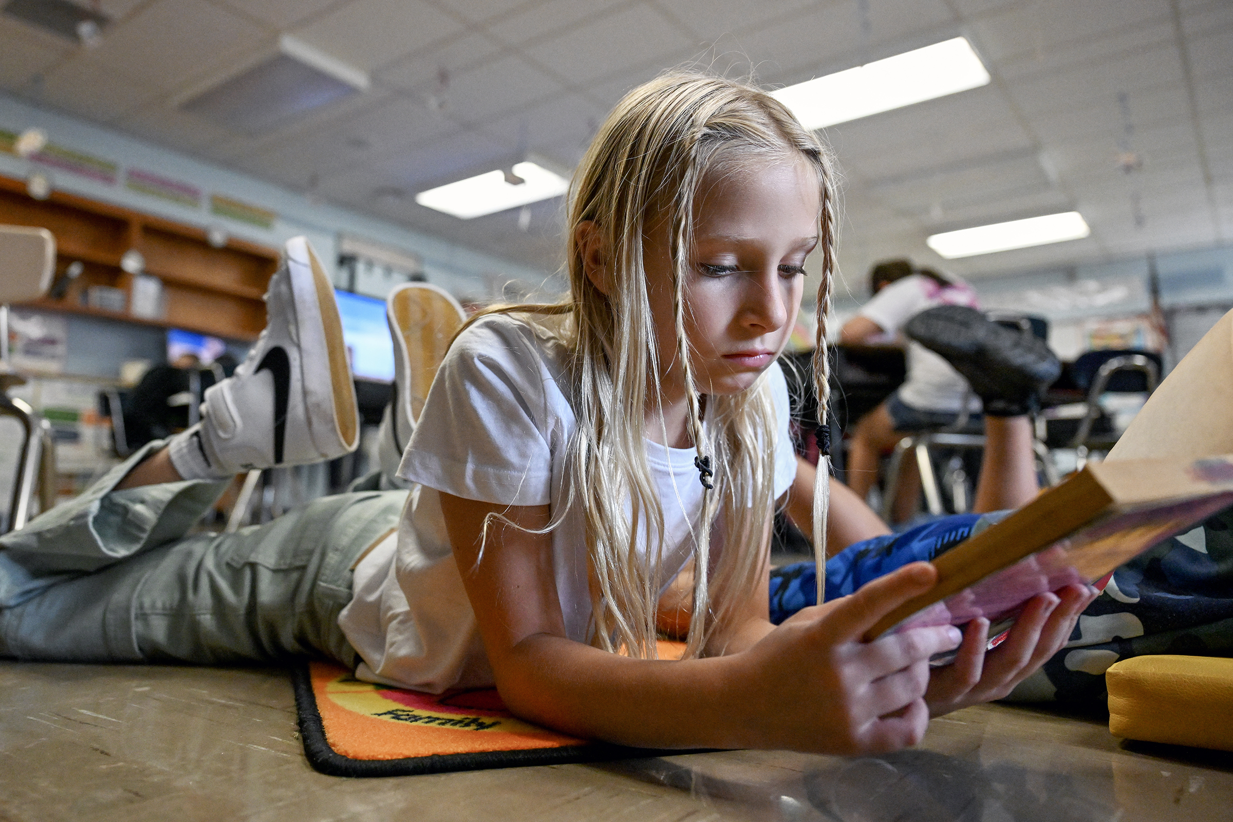 A student lays on her stomach while reading a book in an elementary school library