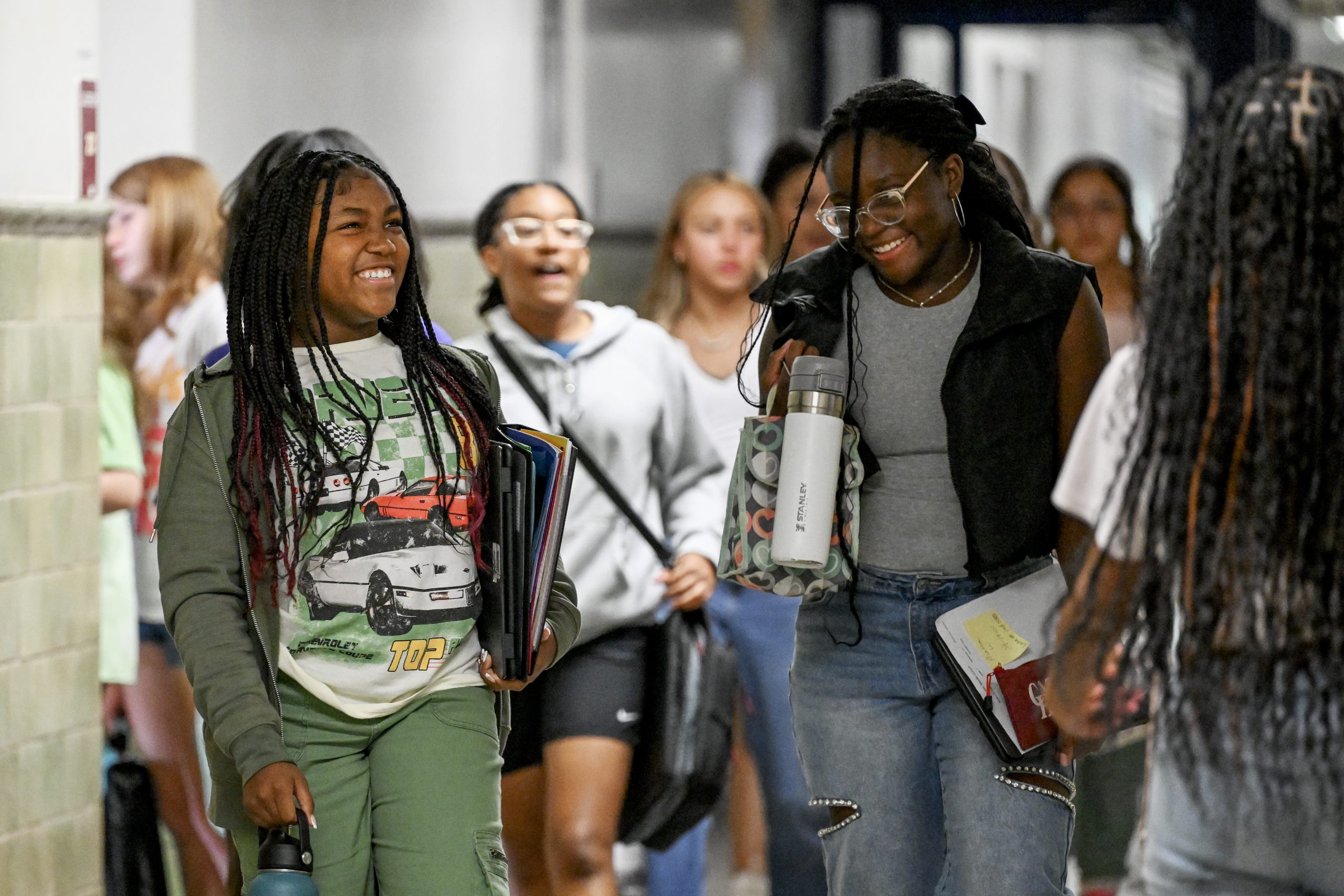 Two teenagers holding books and backpacks walk down a school hallway together, laughing and smiling.