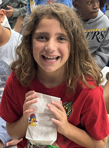 A girl in a red shirt holds a white paper bag with a duck on it. She is smiling.