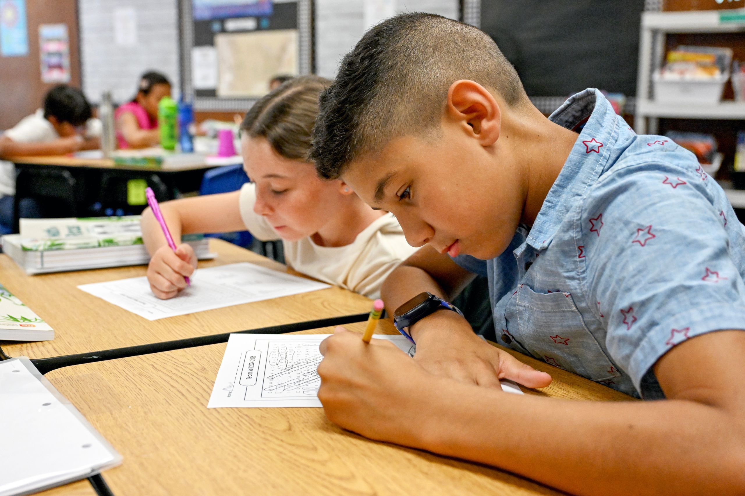 Two students focusing on class work, writing on a paper at their desk.