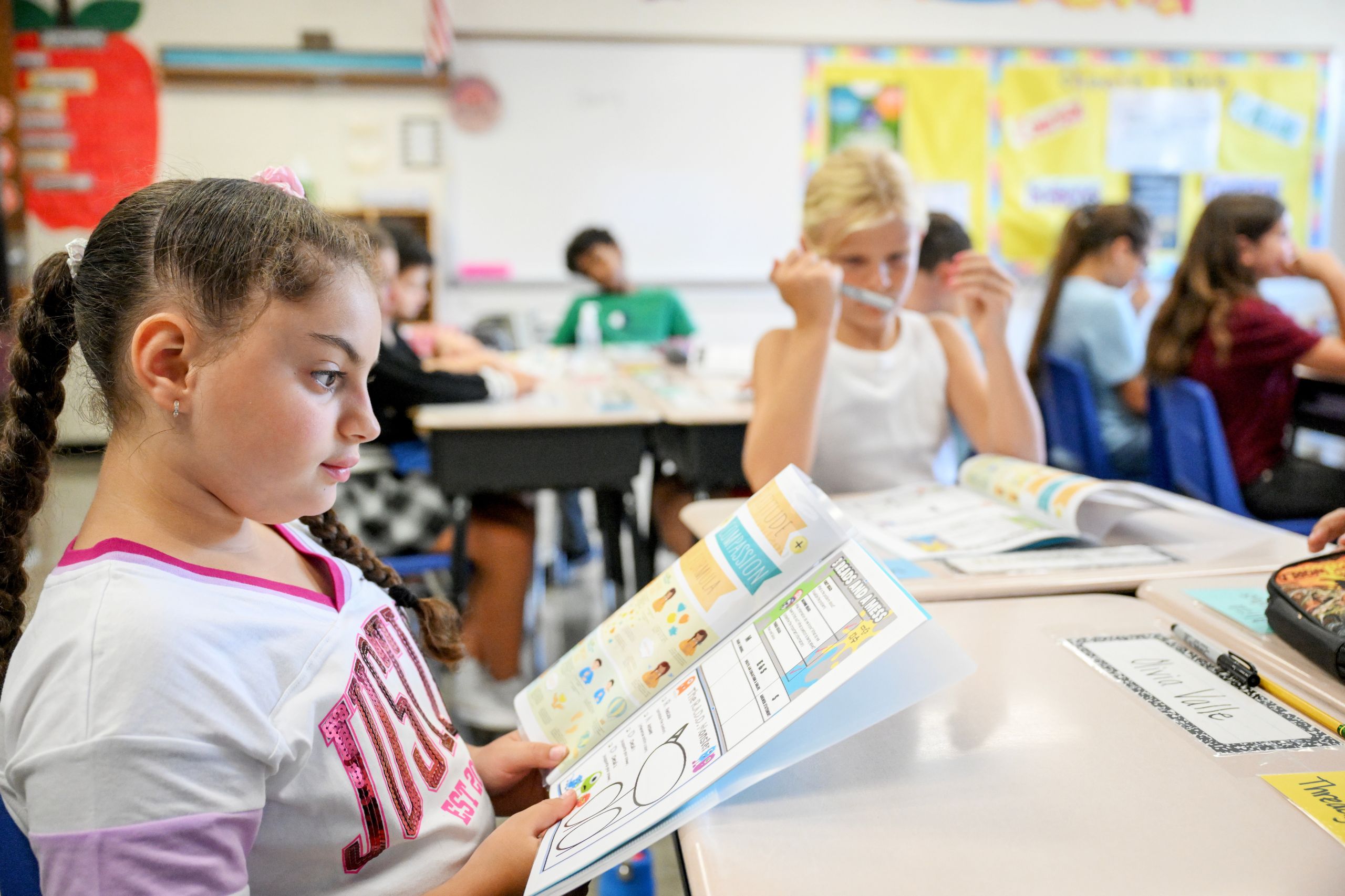 A student holds a book open and reads at their desk with students in the background doing the same.