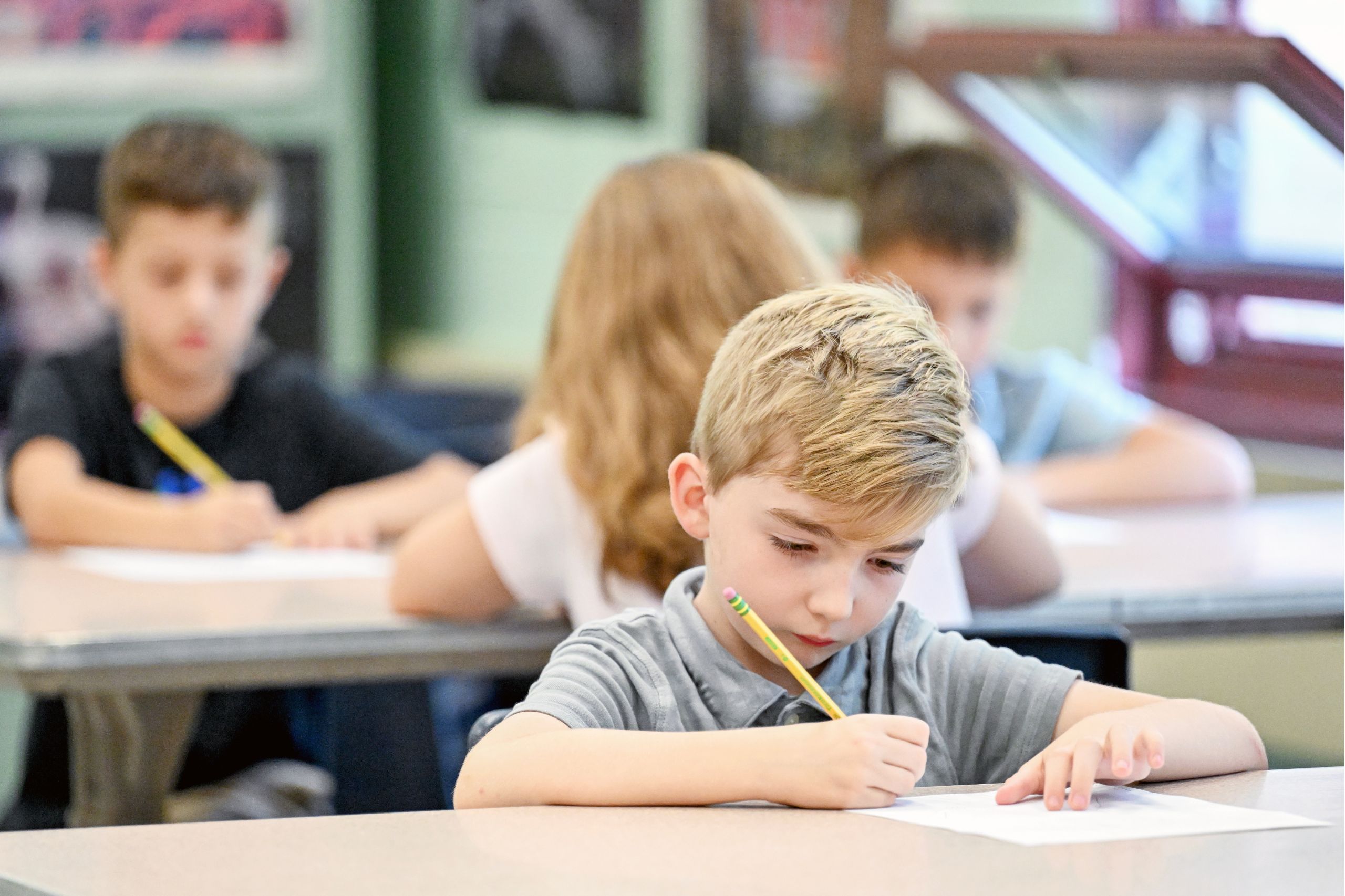 A student is focused on their classwork, writing with a pencil in the air with more students behind them are also focused on class work.