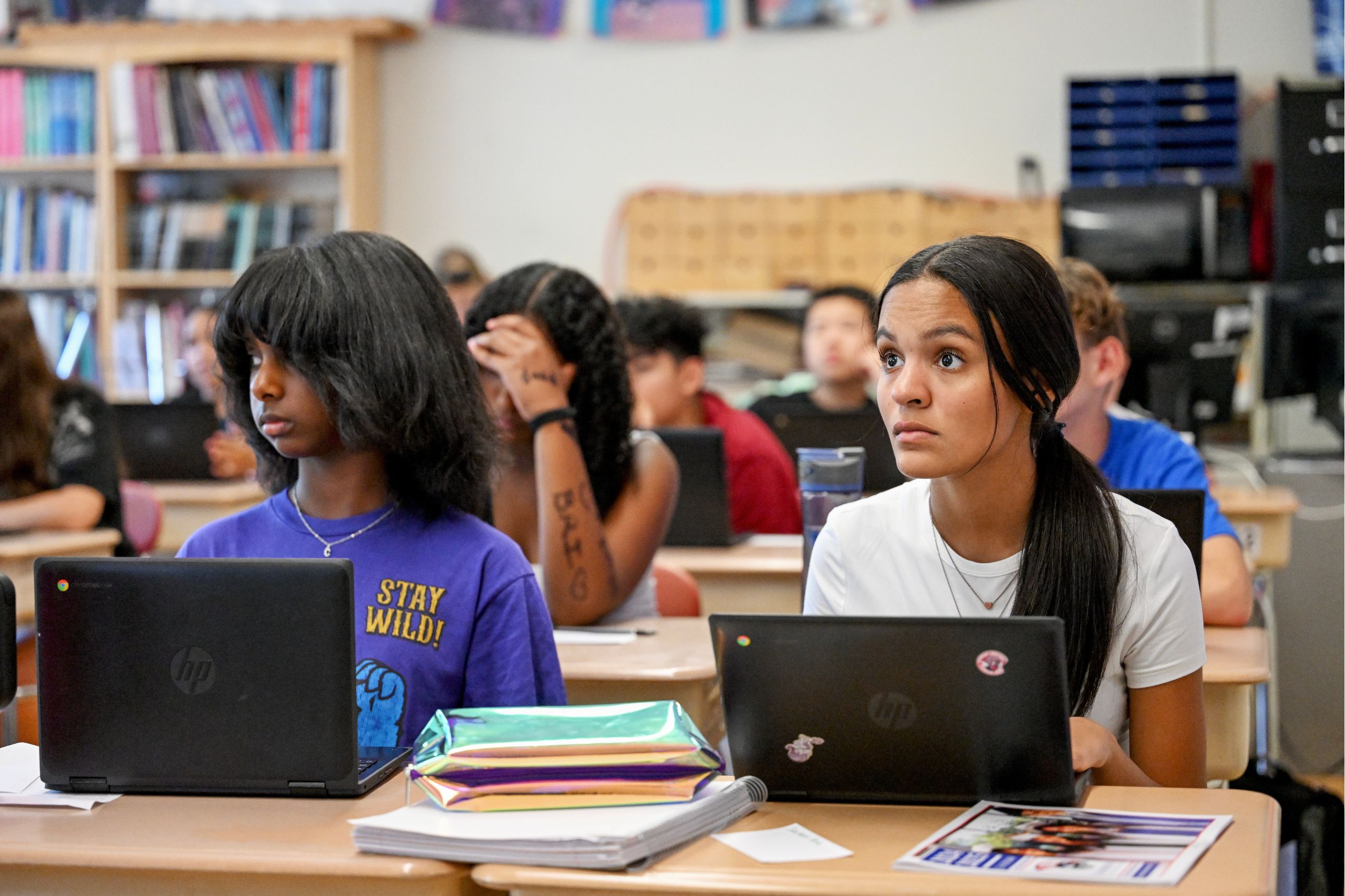 Two students at a desk on laptops, looking to the front of the classroom with more students behind them doing the same.