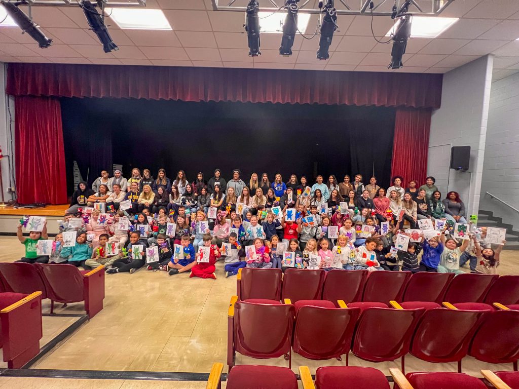 High School and Intermediate School students smile in a group photo with drawings of monsters that the older students sewed together for the younger ones. 