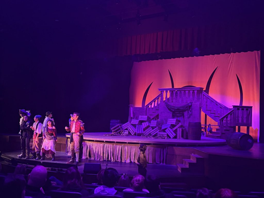 A few students stand in front of the stage at a performance of Treasure Island. The set is dark with a purple glow on the stage with a red background.