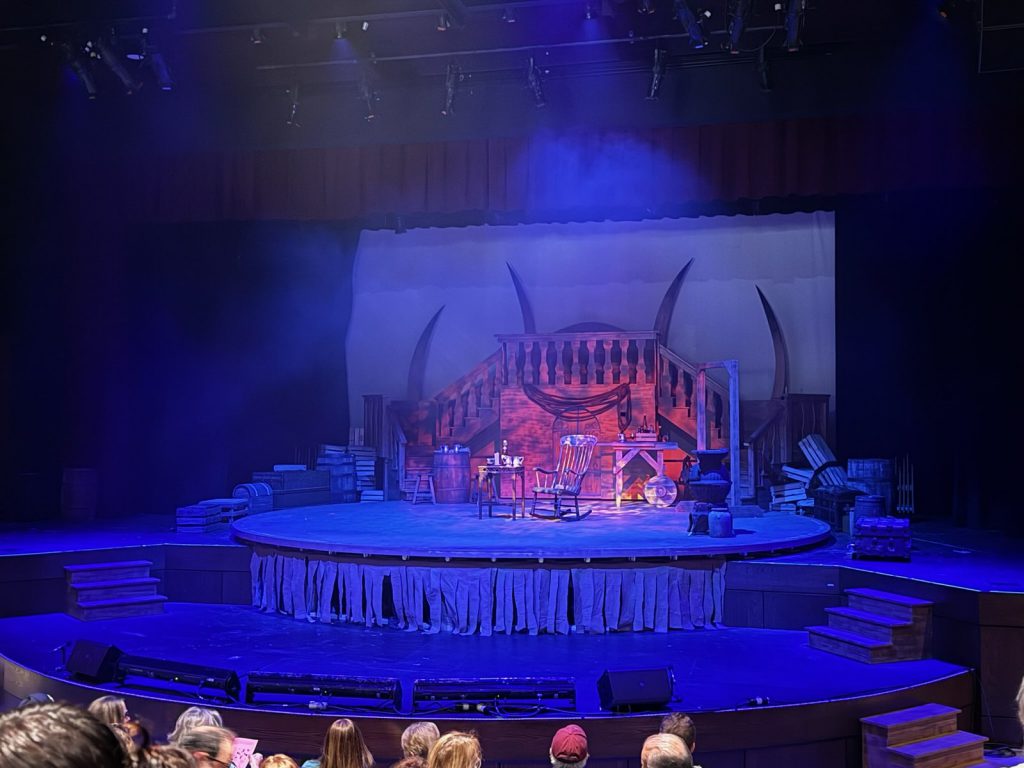 A wide shot of the stage built for the Treasure Island production. The stage is blue with a red light in the center. The picture is from the night of a performance and has family and friends in the audience.