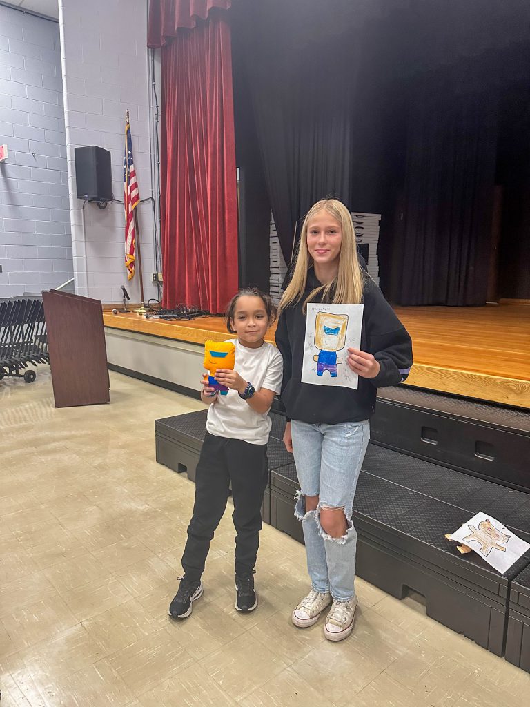 A Goshen High School student and Goshen Intermediate Student pose together. The high school student holds the drawing, and the intermediate student holds the sewn monster they collaborated on. 