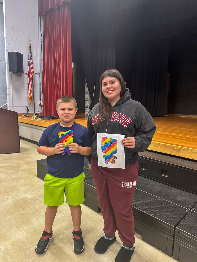 A Goshen High School student and Goshen Intermediate Student pose together. The high school student holds the drawing, and the intermediate student holds the sewn monster they collaborated on. 