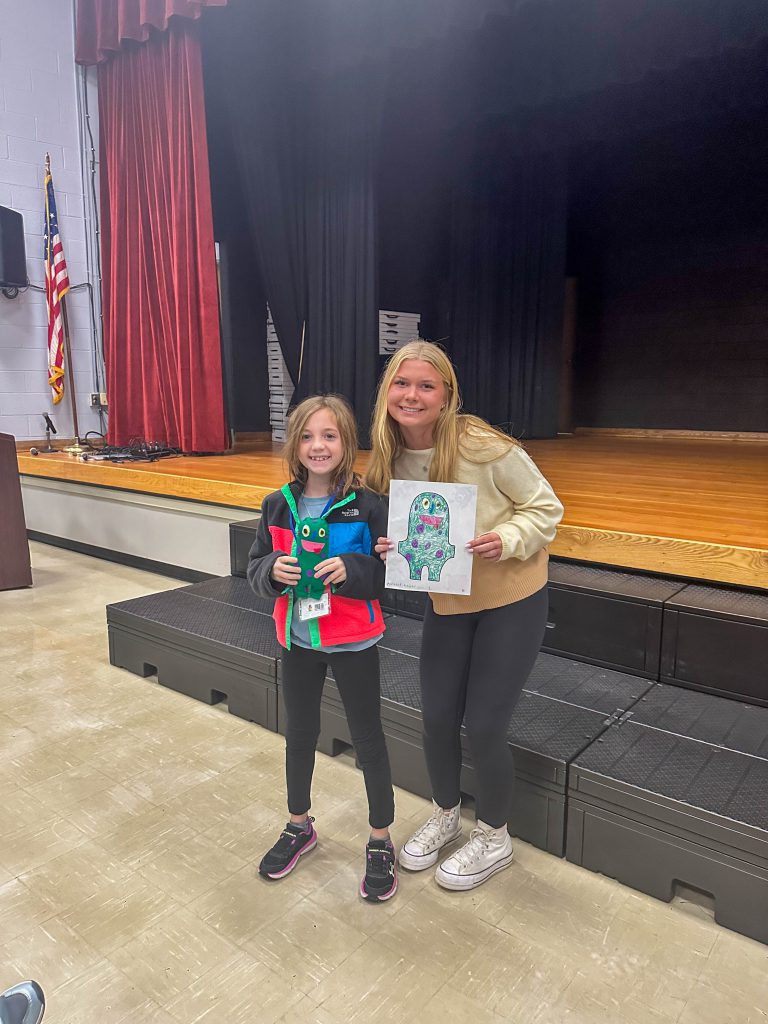 A Goshen High School student and Goshen Intermediate Student pose together. The high school student holds the drawing, and the intermediate student holds the sewn monster they collaborated on. 