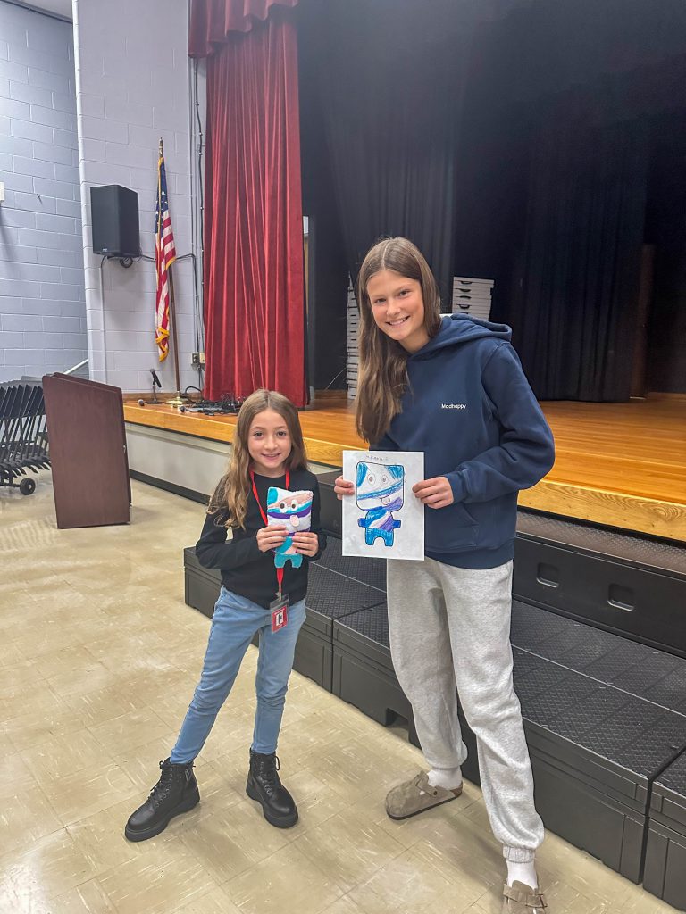 A Goshen High School student and Goshen Intermediate Student pose together. The high school student holds the drawing, and the intermediate student holds the sewn monster they collaborated on. 