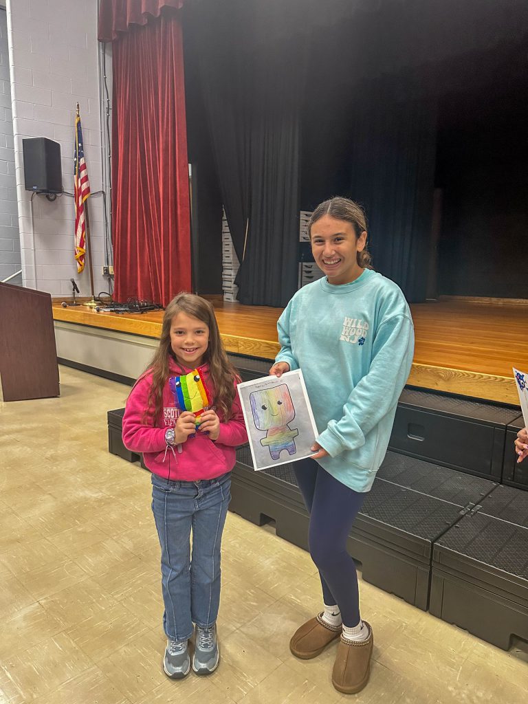 A Goshen High School student and Goshen Intermediate Student pose together. The high school student holds the drawing, and the intermediate student holds the sewn monster they collaborated on. 