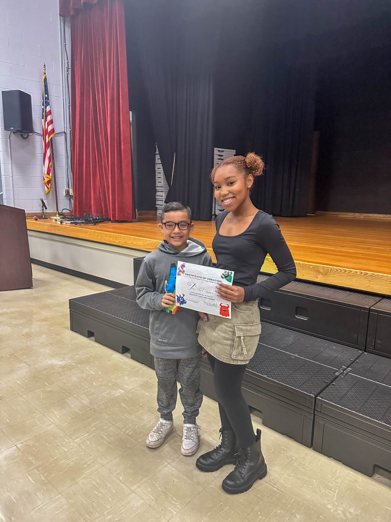 A Goshen High School student and Goshen Intermediate Student pose together. The high school student holds the drawing, and the intermediate student holds the sewn monster they collaborated on. 