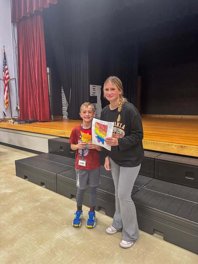 A Goshen High School student and Goshen Intermediate Student pose together. The high school student holds the drawing, and the intermediate student holds the sewn monster they collaborated on. 