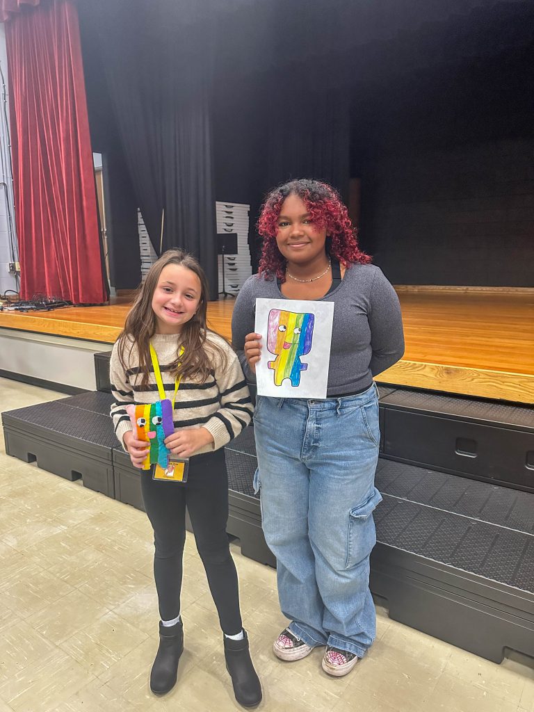 A Goshen High School student and Goshen Intermediate Student pose together. The high school student holds the drawing, and the intermediate student holds the sewn monster they collaborated on. 