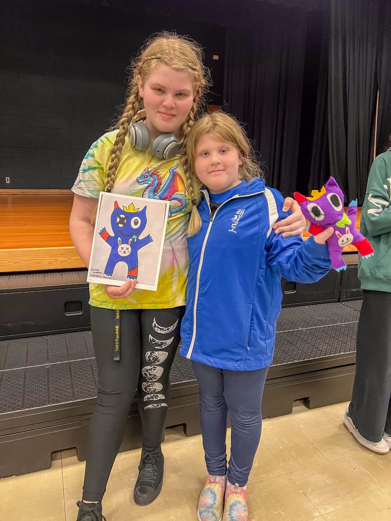 A Goshen High School student and Goshen Intermediate Student pose together. The high school student holds the drawing, and the intermediate student holds the sewn monster they collaborated on. 