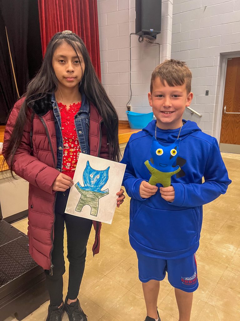 A Goshen High School student and Goshen Intermediate Student pose together. The high school student holds the drawing, and the intermediate student holds the sewn monster they collaborated on. 