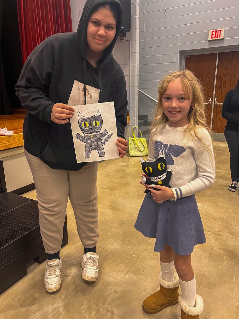 A Goshen High School student and Goshen Intermediate Student pose together. The high school student holds the drawing, and the intermediate student holds the sewn monster they collaborated on. 