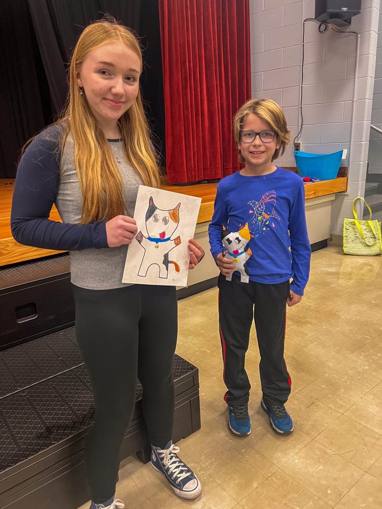 A Goshen High School student and Goshen Intermediate Student pose together. The high school student holds the drawing, and the intermediate student holds the sewn monster they collaborated on. 