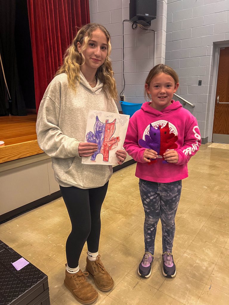 A Goshen High School student and Goshen Intermediate Student pose together. The high school student holds the drawing, and the intermediate student holds the sewn monster they collaborated on. 