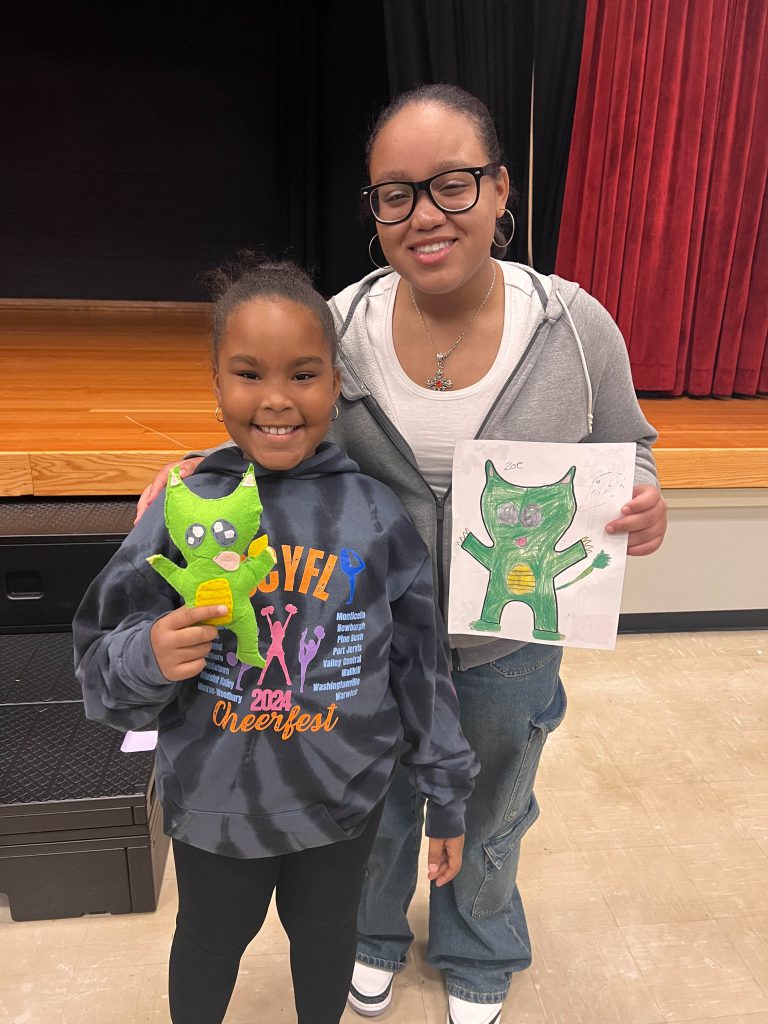 A Goshen High School student and Goshen Intermediate Student pose together. The high school student holds the drawing, and the intermediate student holds the sewn monster they collaborated on. 