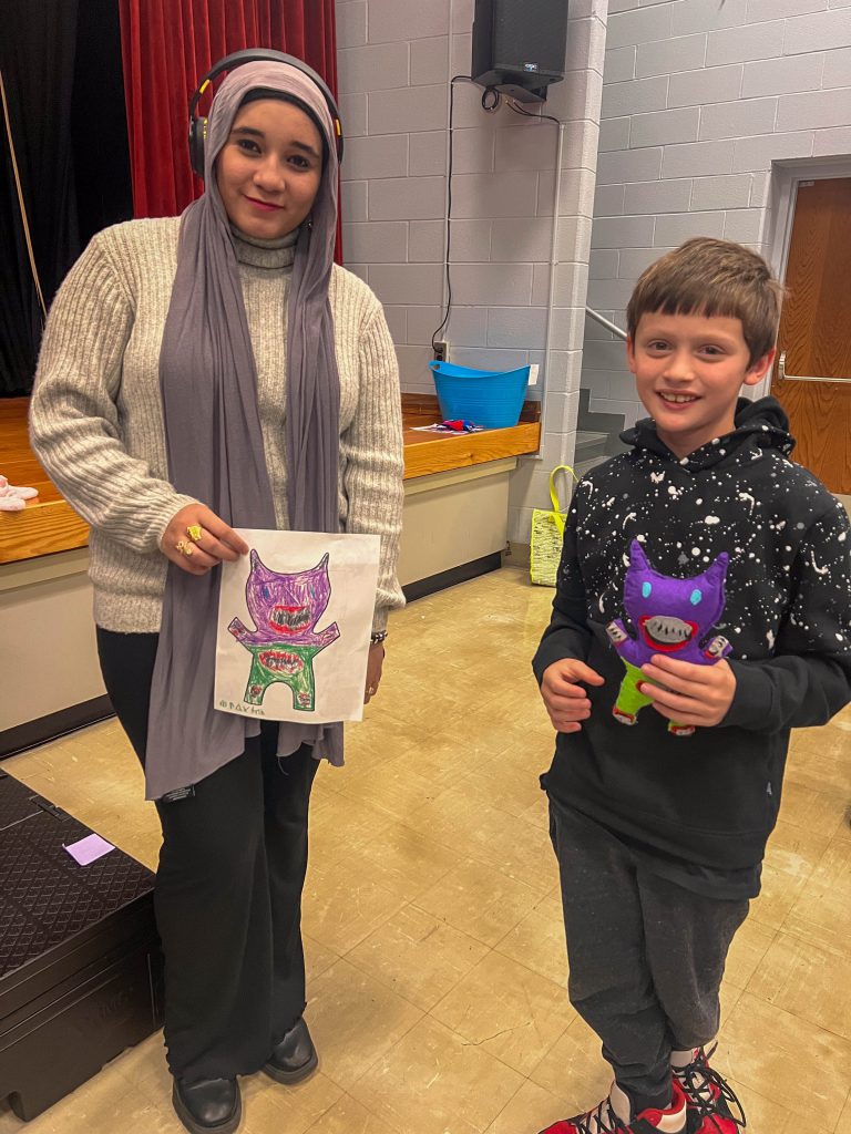 A Goshen High School student and Goshen Intermediate Student pose together. The high school student holds the drawing, and the intermediate student holds the sewn monster they collaborated on. 