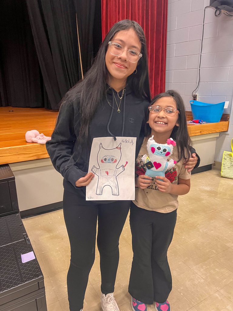 A Goshen High School student and Goshen Intermediate Student pose together. The high school student holds the drawing, and the intermediate student holds the sewn monster they collaborated on. 