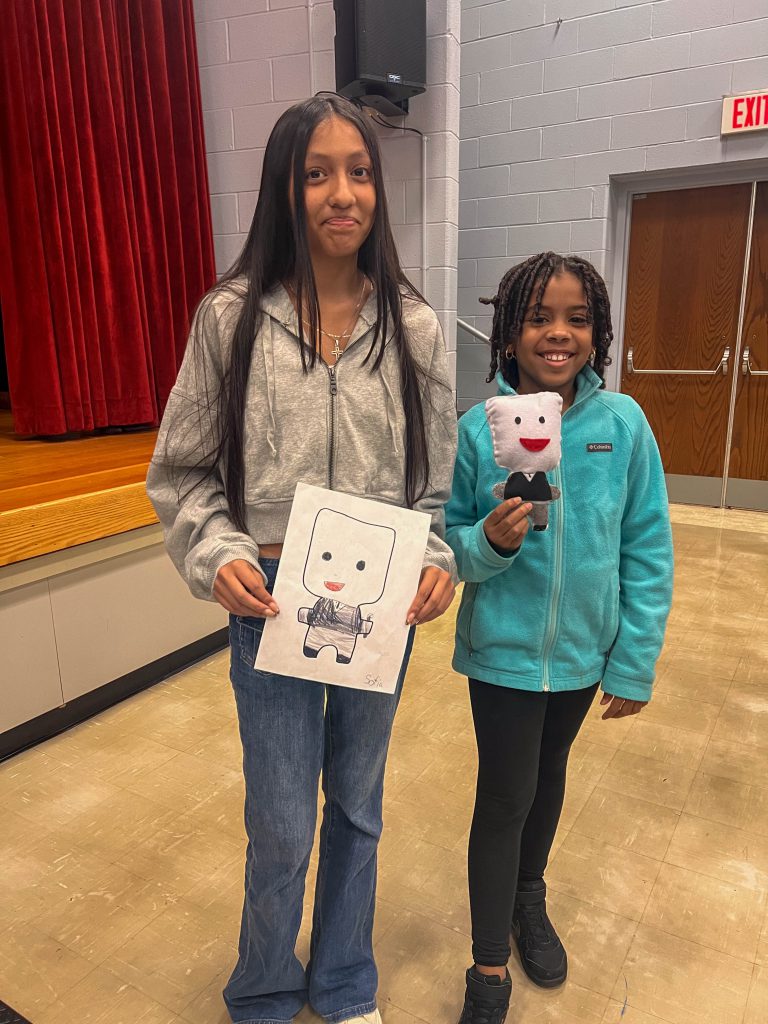 A Goshen High School student and Goshen Intermediate Student pose together. The high school student holds the drawing, and the intermediate student holds the sewn monster they collaborated on. 