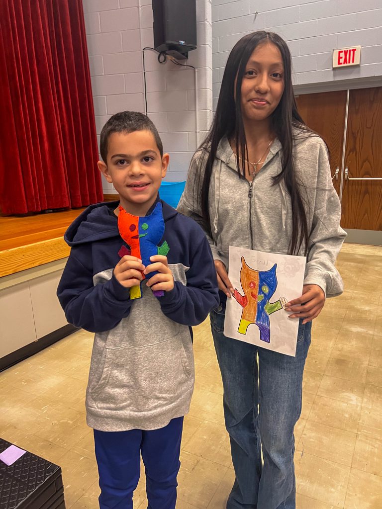 A Goshen High School student and Goshen Intermediate Student pose together. The high school student holds the drawing, and the intermediate student holds the sewn monster they collaborated on. 