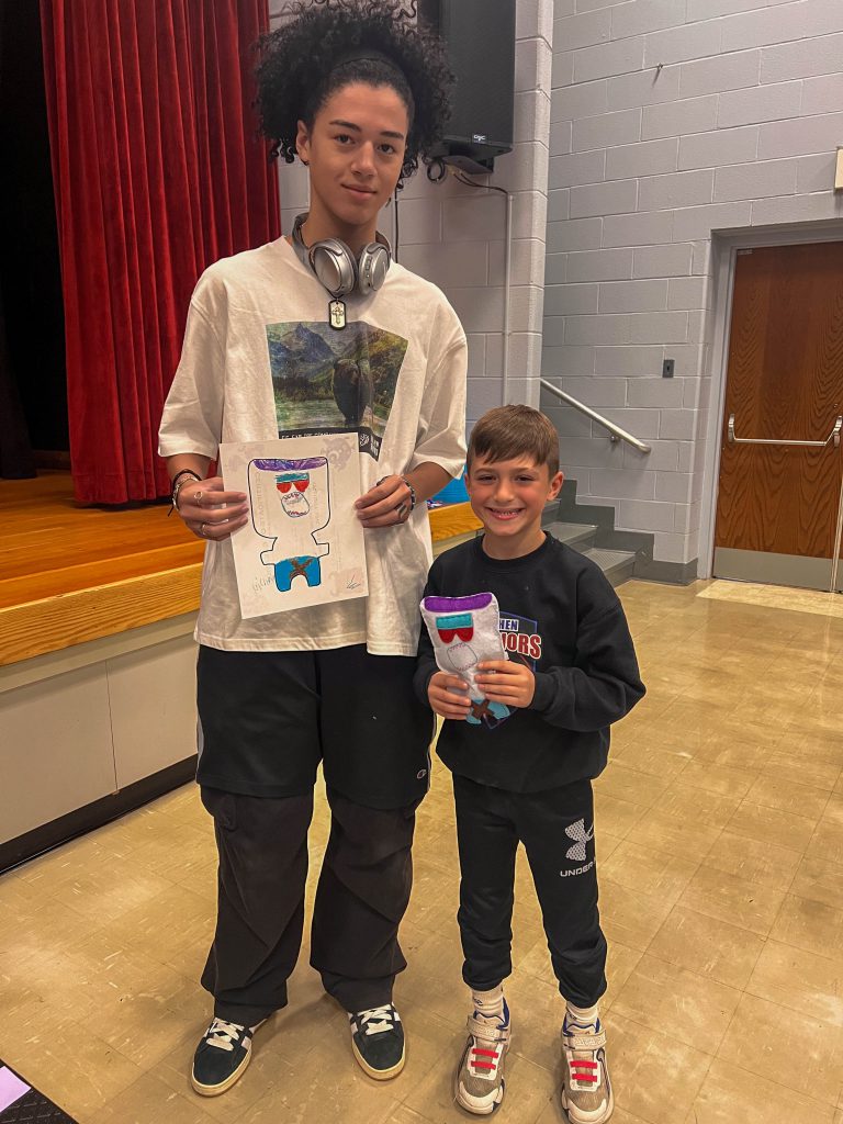 A Goshen High School student and Goshen Intermediate Student pose together. The high school student holds the drawing, and the intermediate student holds the sewn monster they collaborated on. 