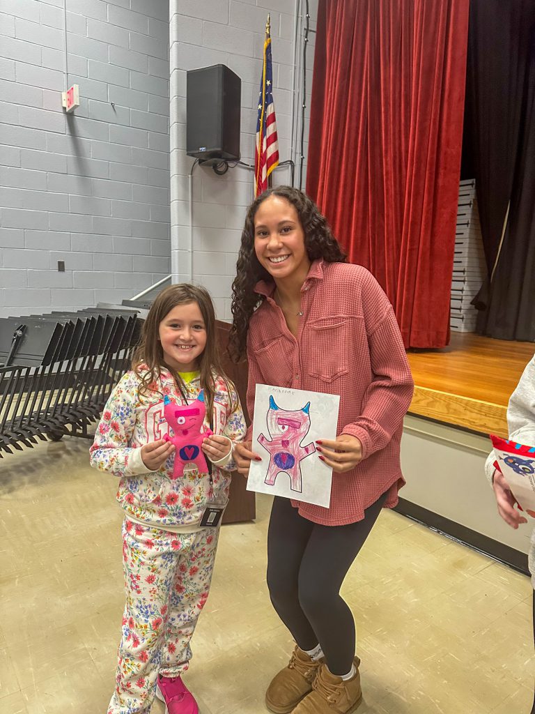 A Goshen High School student and Goshen Intermediate Student pose together. The high school student holds the drawing, and the intermediate student holds the sewn monster they collaborated on. 