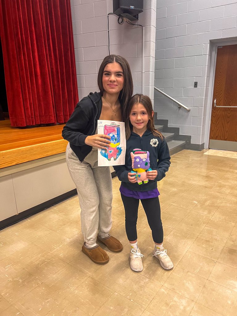 A Goshen High School student and Goshen Intermediate Student pose together. The high school student holds the drawing, and the intermediate student holds the sewn monster they collaborated on. 