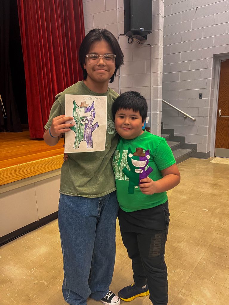 A Goshen High School student and Goshen Intermediate Student pose together. The high school student holds the drawing, and the intermediate student holds the sewn monster they collaborated on. 