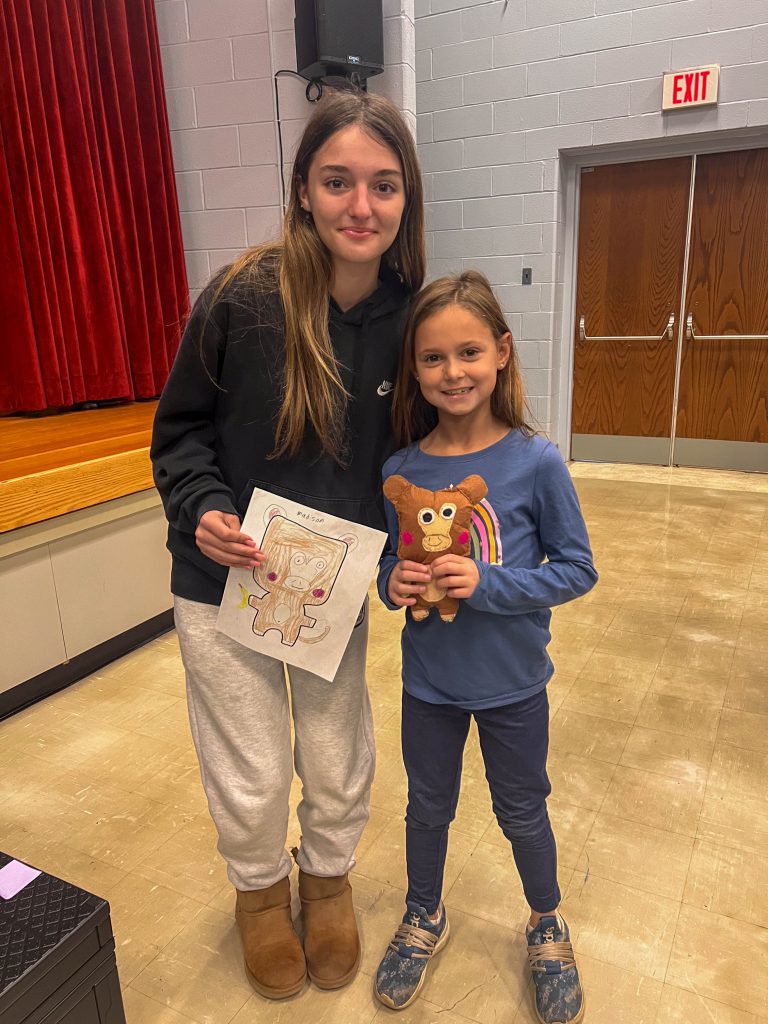 A Goshen High School student and Goshen Intermediate Student pose together. The high school student holds the drawing, and the intermediate student holds the sewn monster they collaborated on. 