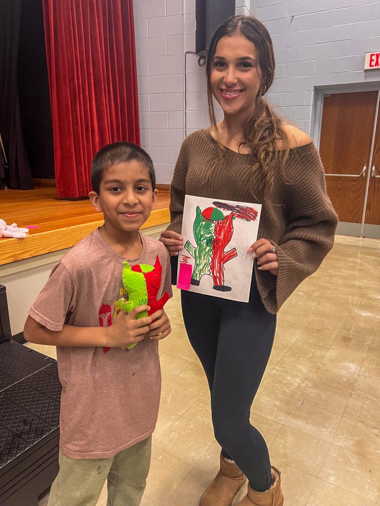 A Goshen High School student and Goshen Intermediate Student pose together. The high school student holds the drawing, and the intermediate student holds the sewn monster they collaborated on. 