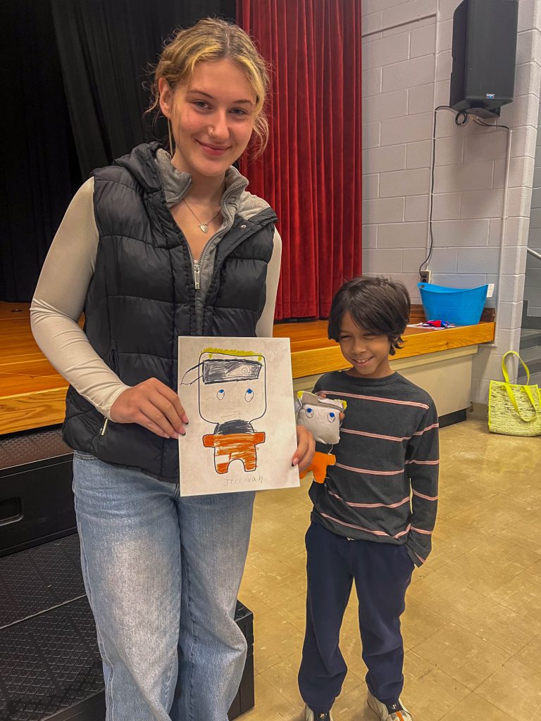 A Goshen High School student and Goshen Intermediate Student pose together. The high school student holds the drawing, and the intermediate student holds the sewn monster they collaborated on. 