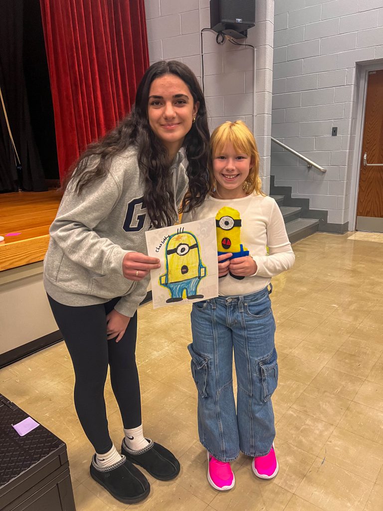 A Goshen High School student and Goshen Intermediate Student pose together. The high school student holds the drawing, and the intermediate student holds the sewn monster they collaborated on. 