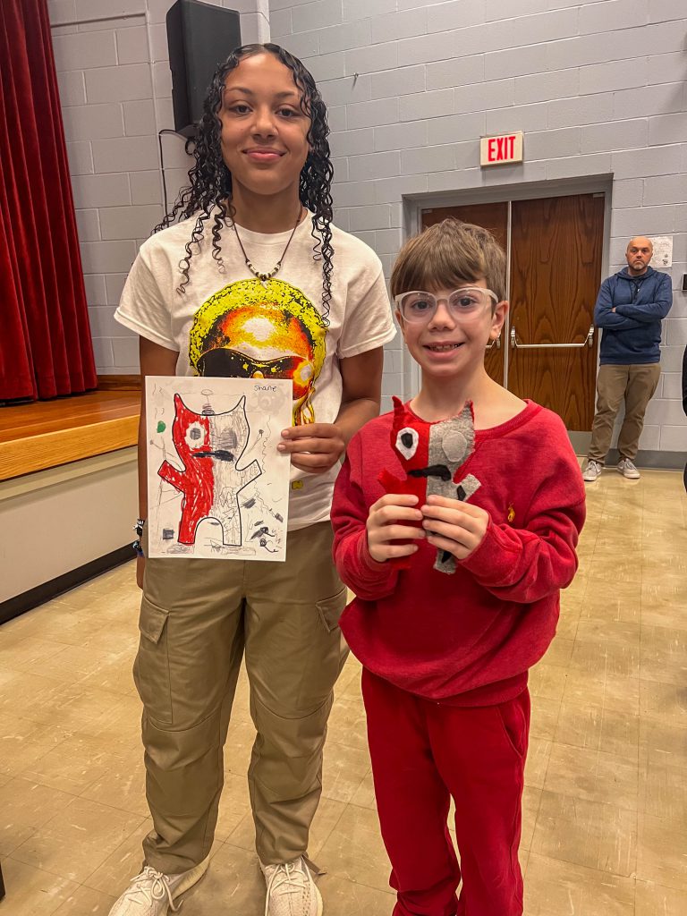 A Goshen High School student and Goshen Intermediate Student pose together. The high school student holds the drawing, and the intermediate student holds the sewn monster they collaborated on. 