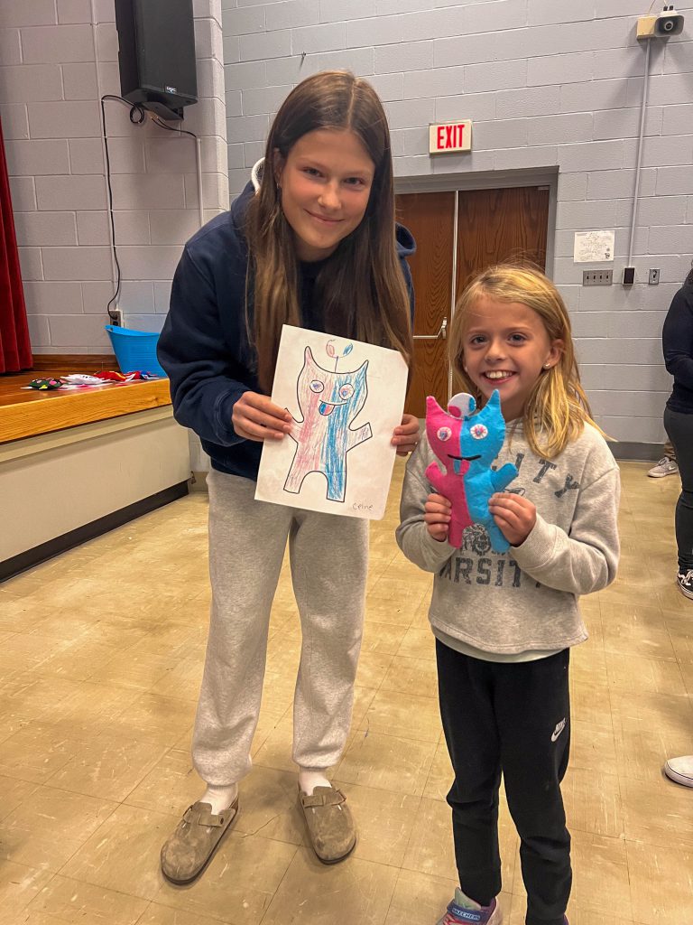 A Goshen High School student and Goshen Intermediate Student pose together. The high school student holds the drawing, and the intermediate student holds the sewn monster they collaborated on. 