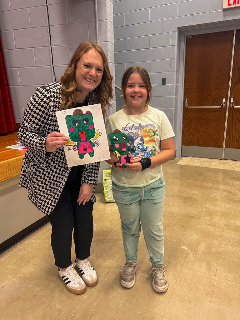 A Goshen teacher and Goshen Intermediate Student pose together. The teacher holds the drawing, and the intermediate student holds the sewn monster they collaborated on.