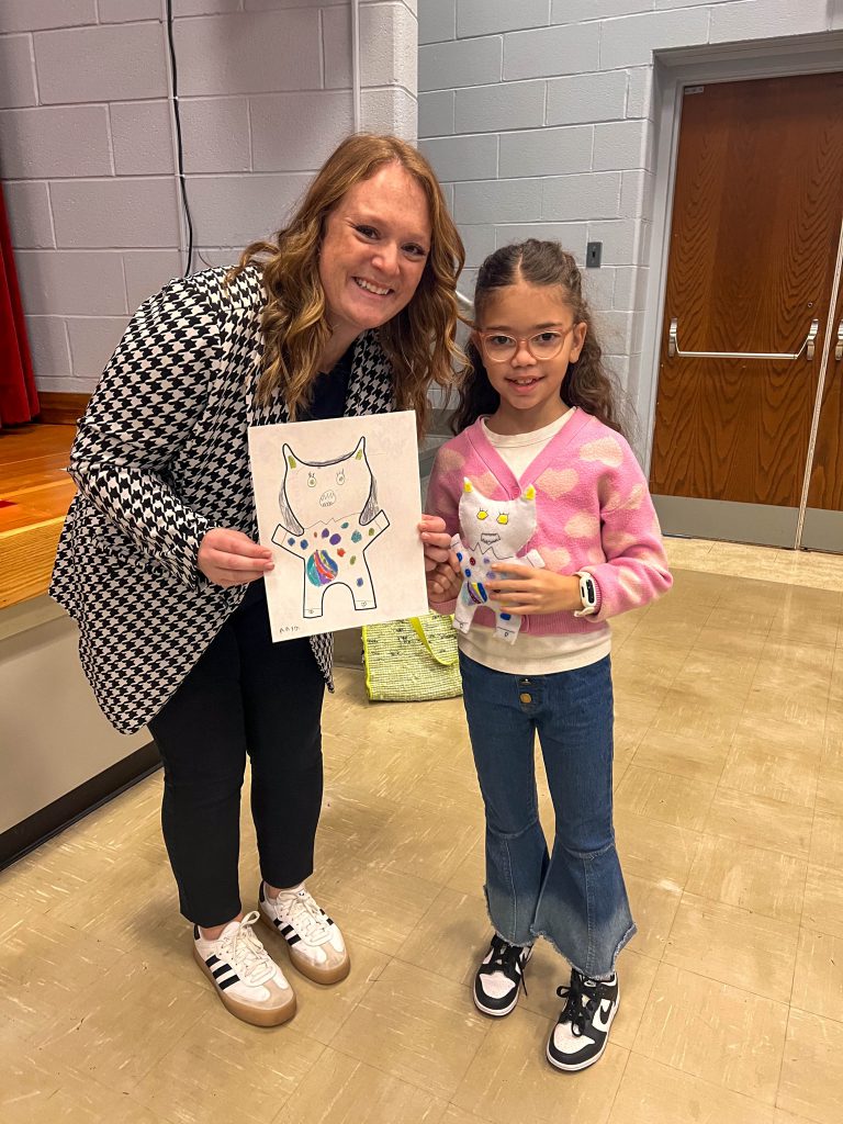 A Goshen teacher and Goshen Intermediate Student pose together. The teacher holds the drawing, and the intermediate student holds the sewn monster they collaborated on.