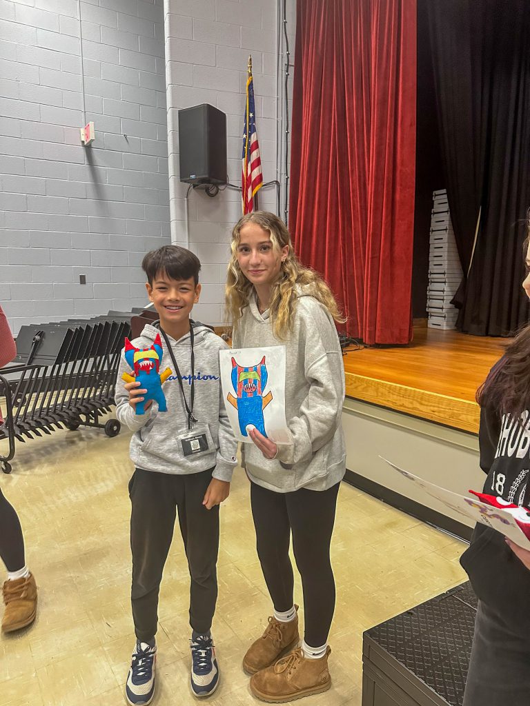 A Goshen High School student and Goshen Intermediate Student pose together. The high school student holds the drawing, and the intermediate student holds the sewn monster they collaborated on. 