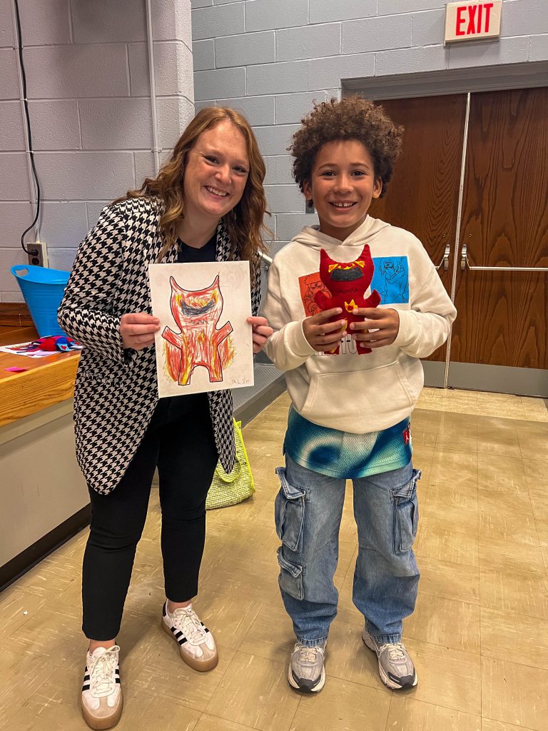 A Goshen teacher and Goshen Intermediate Student pose together. The teacher holds the drawing, and the intermediate student holds the sewn monster they collaborated on. 