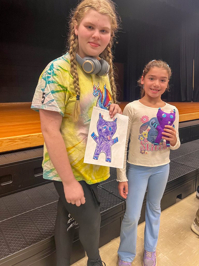 A Goshen High School student and Goshen Intermediate Student pose together. The high school student holds the drawing, and the intermediate student holds the sewn monster they collaborated on. 