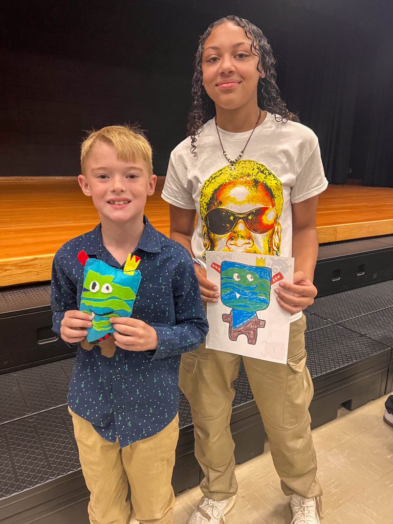 A Goshen High School student and Goshen Intermediate Student pose together. The high school student holds the drawing, and the intermediate student holds the sewn monster they collaborated on. 