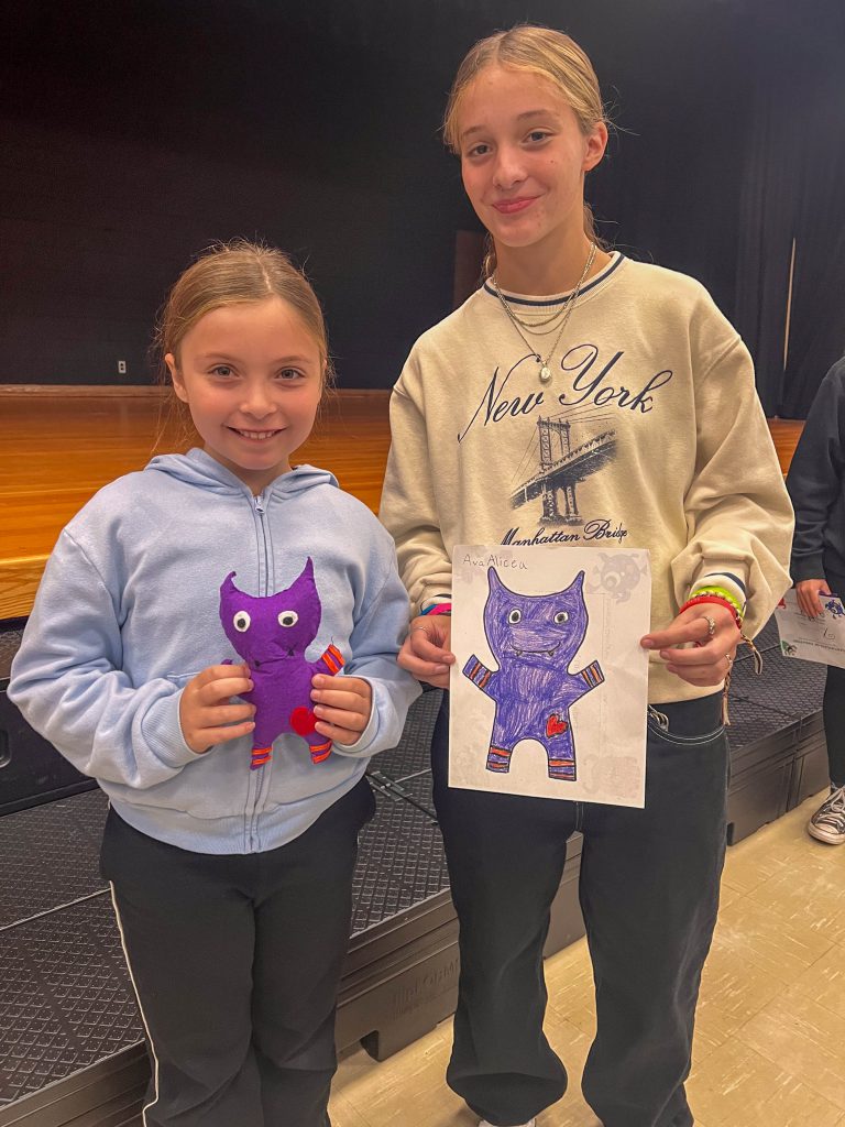 A Goshen High School student and Goshen Intermediate Student pose together. The high school student holds the drawing, and the intermediate student holds the sewn monster they collaborated on. 