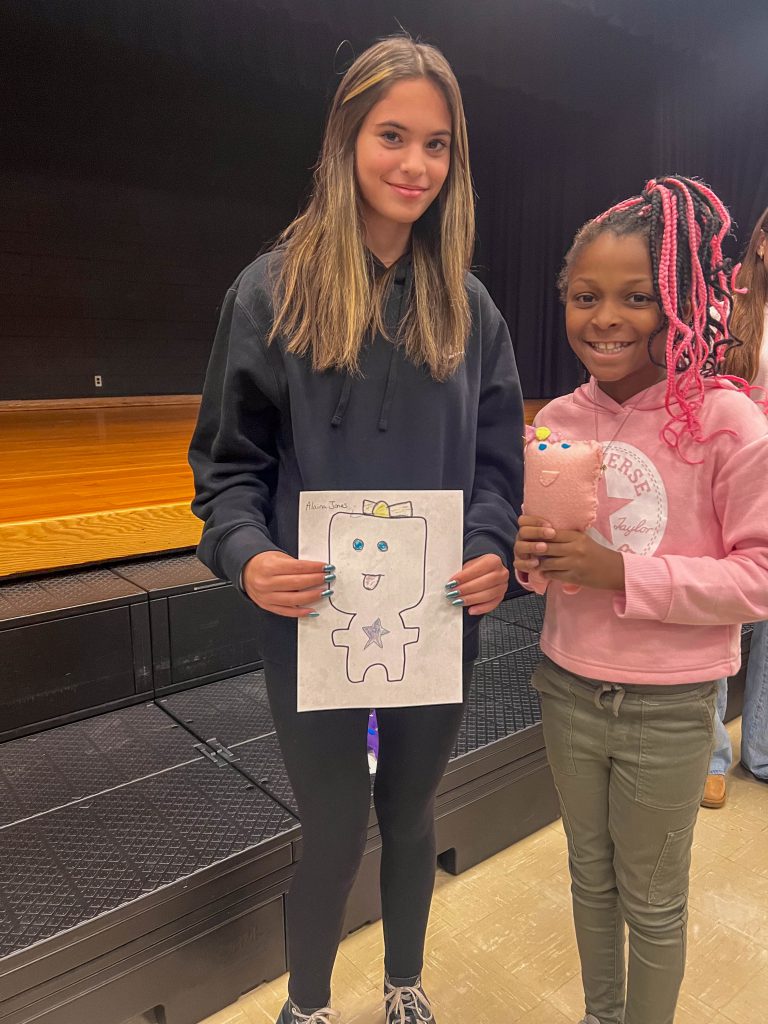 A Goshen High School student and Goshen Intermediate Student pose together. The high school student holds the drawing, and the intermediate student holds the sewn monster they collaborated on. 