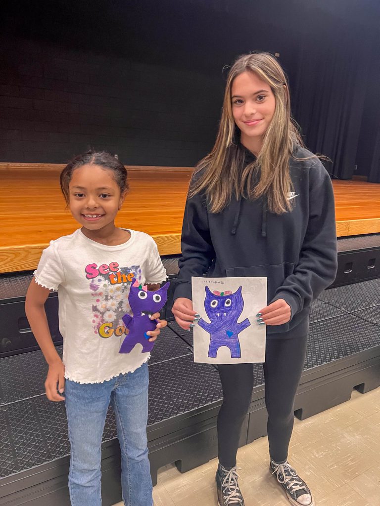 A Goshen High School student and Goshen Intermediate Student pose together. The high school student holds the drawing, and the intermediate student holds the sewn monster they collaborated on. 