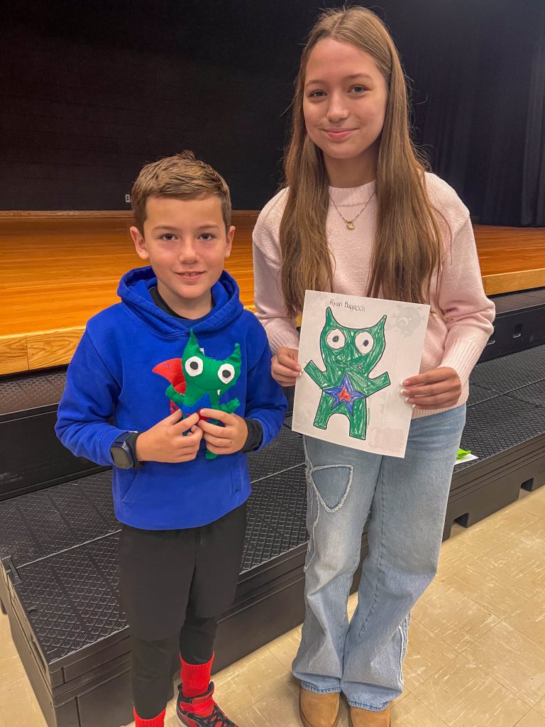 A Goshen High School student and Goshen Intermediate Student pose together. The high school student holds the drawing, and the intermediate student holds the sewn monster they collaborated on. 