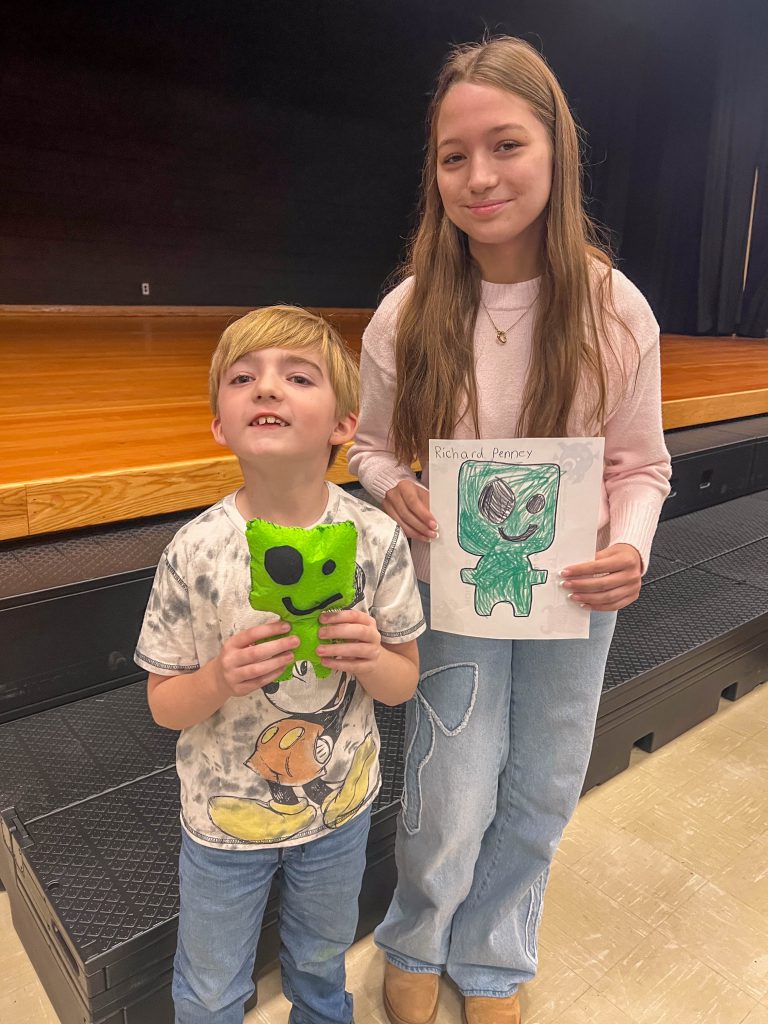 A Goshen High School student and Goshen Intermediate Student pose together. The high school student holds the drawing, and the intermediate student holds the sewn monster they collaborated on. 