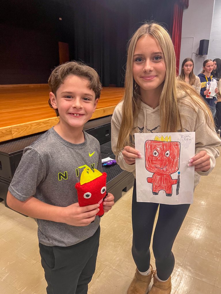 A Goshen High School student and Goshen Intermediate Student pose together. The high school student holds the drawing, and the intermediate student holds the sewn monster they collaborated on. 